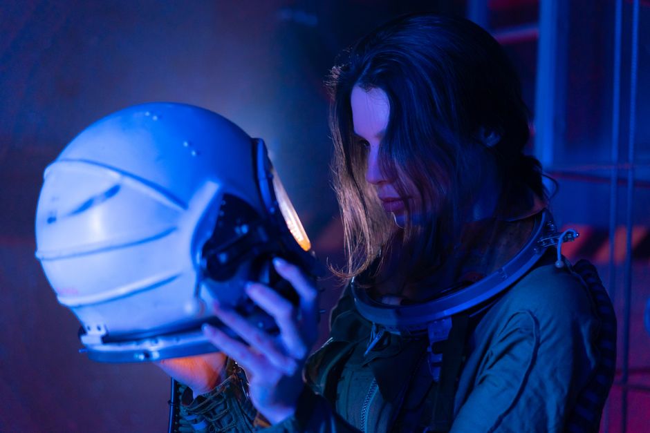 Una mujer en un traje espacial examina un casco de astronauta bajo una iluminación futurista azul.