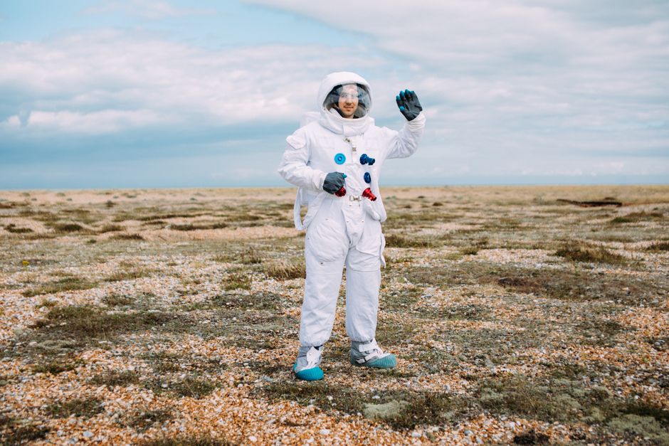 Astronauta con un traje espacial saludando en un paisaje desolado y abierto bajo un cielo nublado.