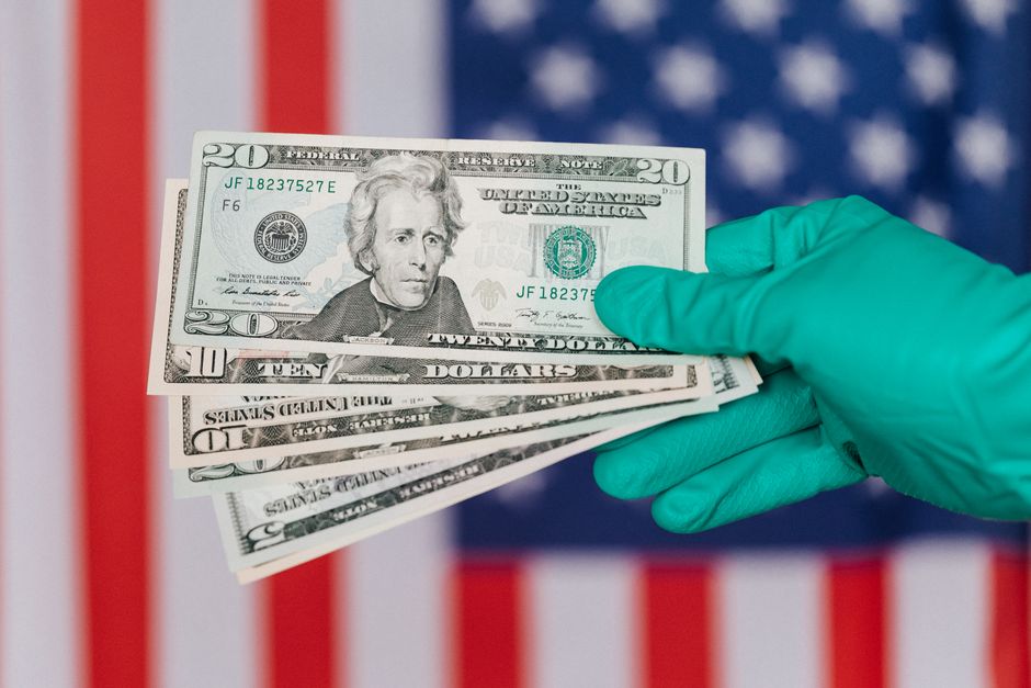 Una mano enguantada sostiene billetes de dólar estadounidenses frente a una bandera americana, simbolizando finanzas y protección.