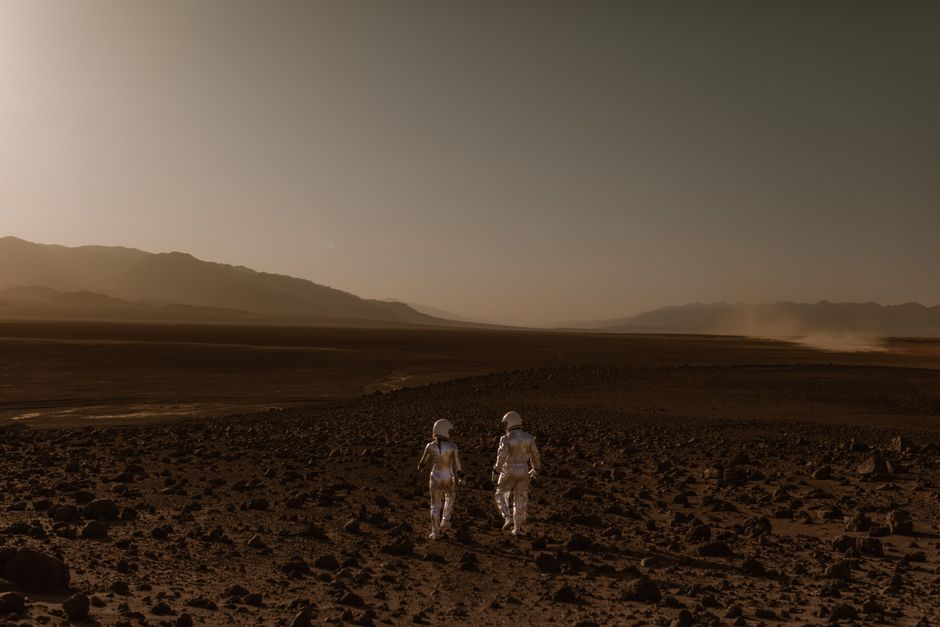 Dos astronautas con trajes espaciales caminan por un paisaje desértico similar a Marte durante el día.