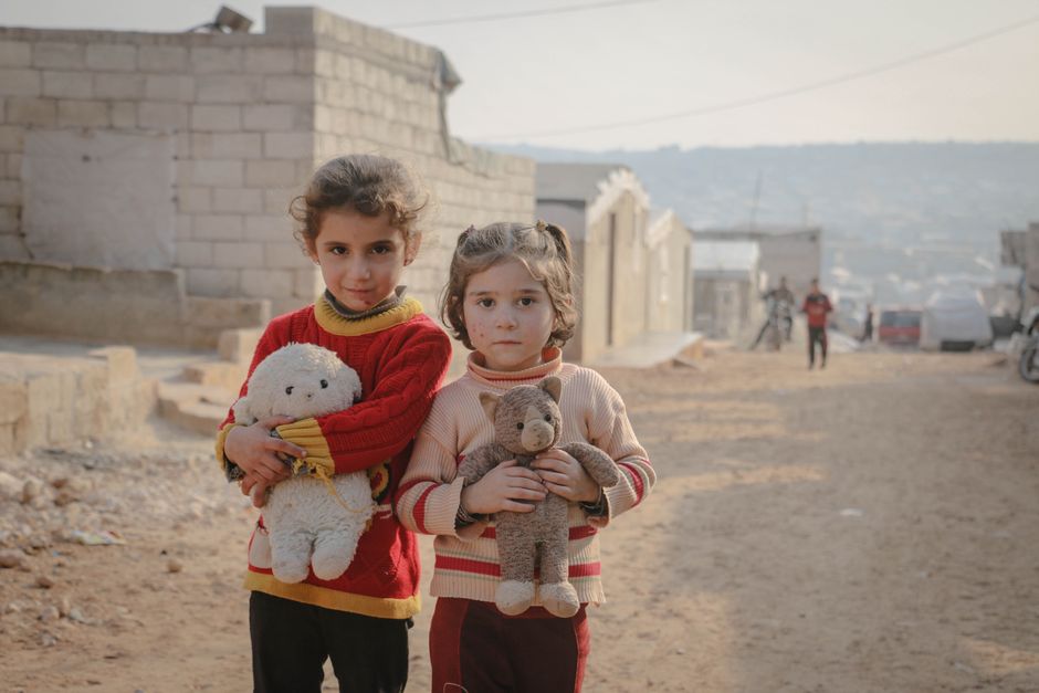 Dos niñas pequeñas con juguetes de peluche en una calle de Idlib, Siria, capturando resiliencia e inocencia.