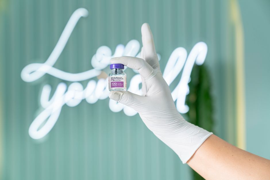 Una mano enguantada sostiene un vial de Botox frente a un suave fondo verde con luces de neón.