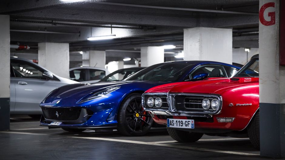 Un Ferrari azul y un Pontiac Firebird rojo estacionados en un garaje subterráneo.
