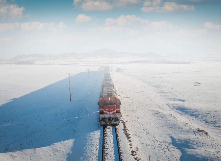 Un tren rojo solitario viaja a través de un vasto paisaje nevado bajo un cielo brillante.