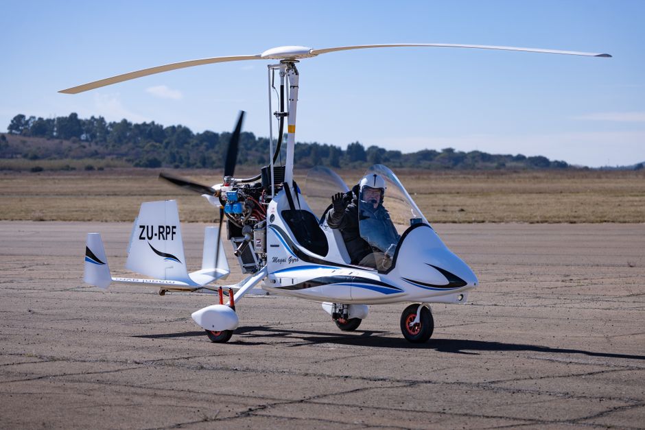 Un piloto saluda desde un autogiro blanco estacionado en una soleada pista rural.