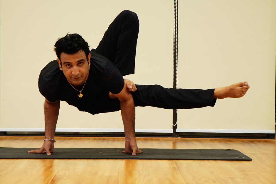 Un hombre adulto se equilibra en la pose de libélula sobre una esterilla de yoga en interiores, representando fuerza y concentración.