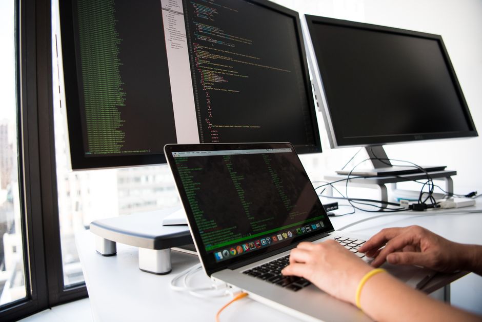 Un desarrollador escribe código en un laptop frente a múltiples monitores en un entorno de oficina.