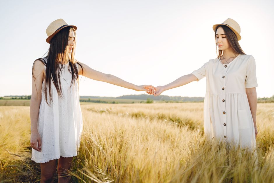 Dos jóvenes mujeres de la mano en un soleado campo de trigo, vistiendo vestidos blancos y sombreros.