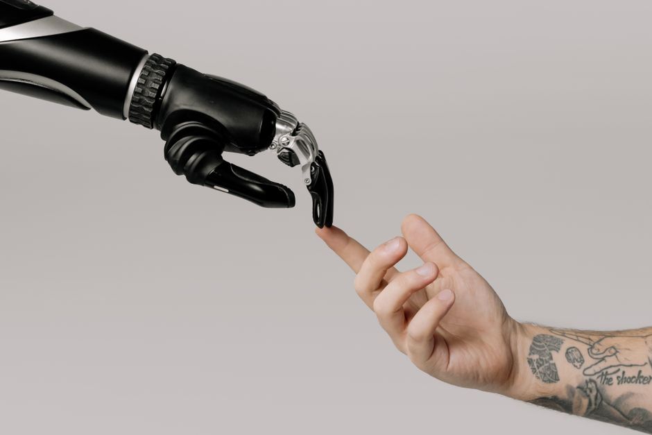 Una mano humana con tatuajes extendiéndose hacia una mano robótica sobre un fondo blanco.