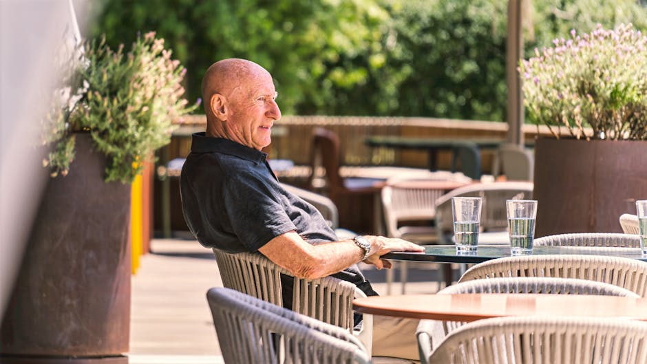Hombre mayor sentado en un patio al aire libre, disfrutando de un día soleado con bebidas en la mesa.