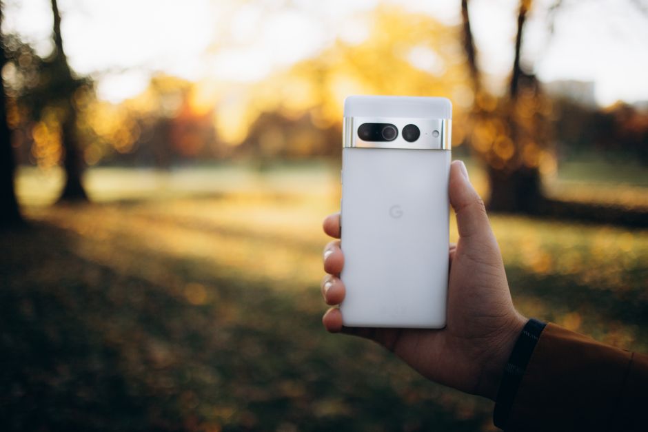 Mano sosteniendo un smartphone blanco frente a un fondo de parque otoñal, capturando la naturaleza y la tecnología.
