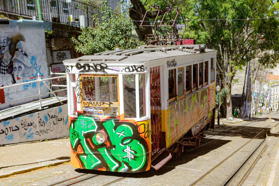 Un tranvía iluminado por el sol cubierto de grafitis recorre una colorida calle urbana, rodeado de arte y árboles.