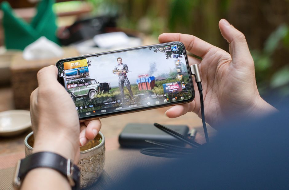 Persona jugando a PUBG en un smartphone al aire libre con un cable de carga conectado.