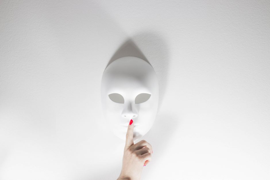 Una máscara blanca con una mano humana gesticulando silencio sobre un fondo blanco brillante, creando una atmósfera misteriosa.