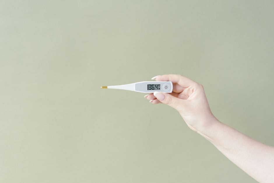 Mano sosteniendo un termómetro digital que muestra 36,4°C sobre un fondo verde claro, estilo minimalista.