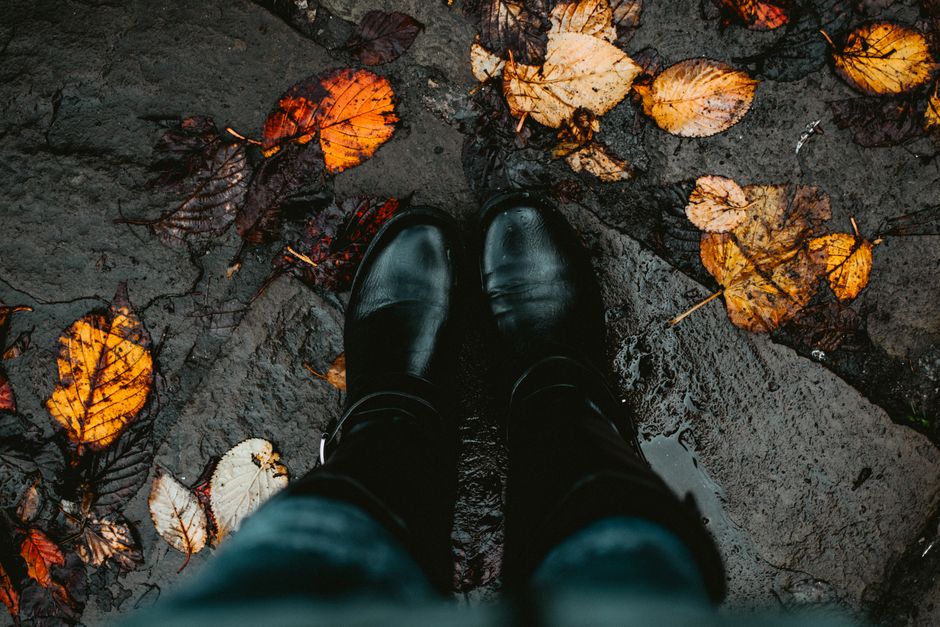 Dos botas negras en un camino de piedra mojada cubierto de hojas, capturando la esencia del otoño.