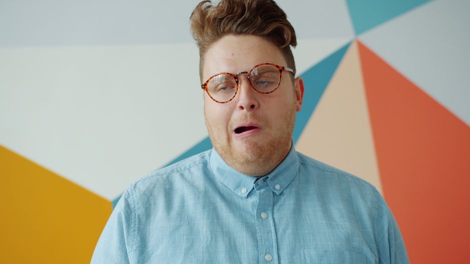 Un joven con gafas hace una expresión humorística frente a una pared geométrica colorida.