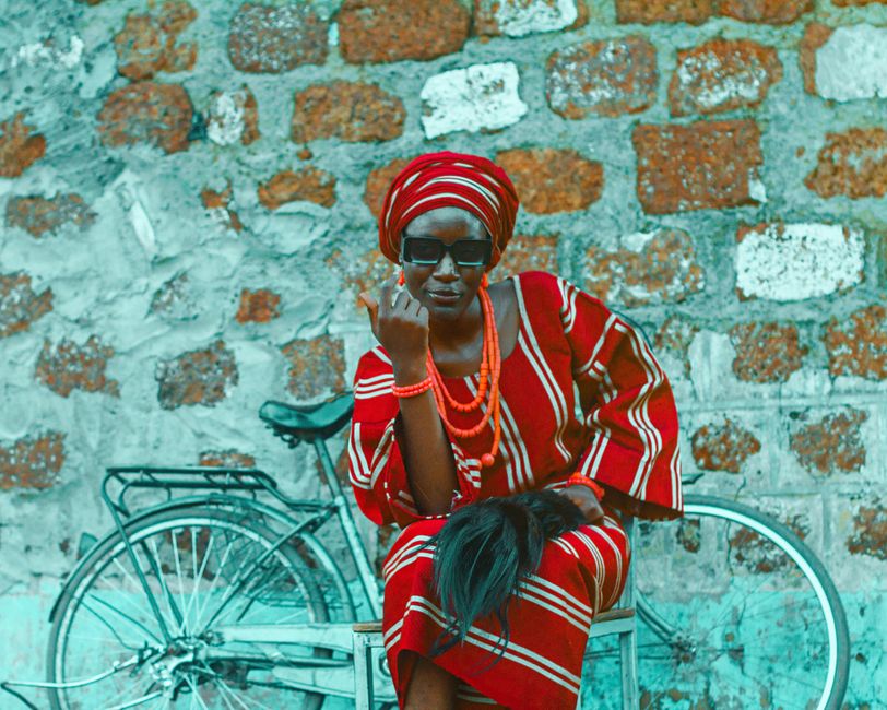 Mujer africana con un llamativo atuendo tradicional, sentada junto a una bicicleta apoyada contra una pared rústica.