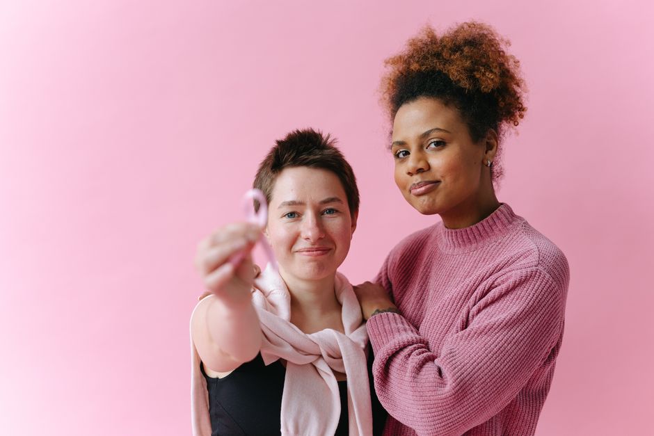 Dos mujeres abrazándose, una sosteniendo una cinta rosa para la concienciación sobre el cáncer de mama, frente a un fondo rosa.
