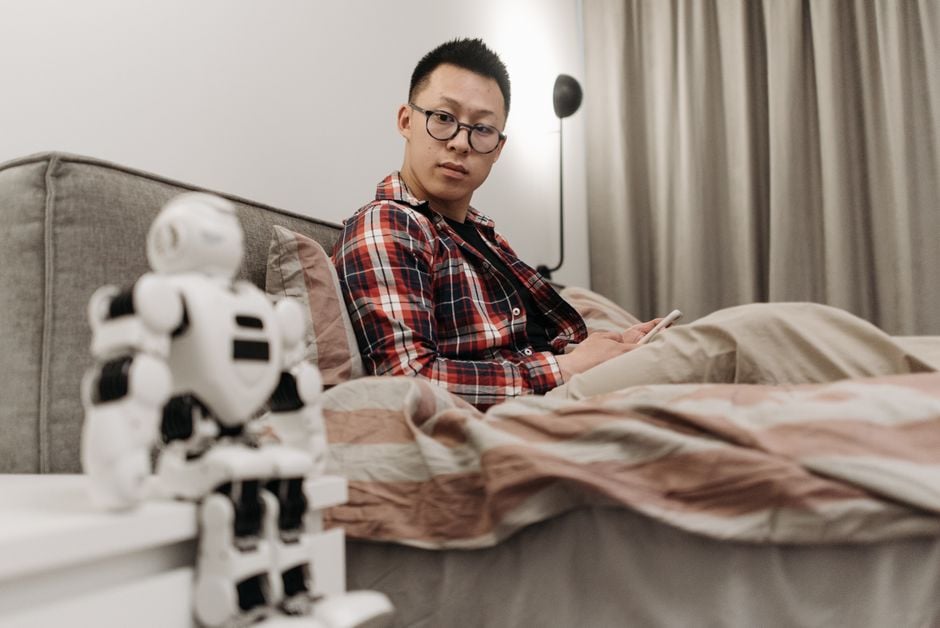 Hombre asiático con camisa de cuadros sentado en la cama, observando un juguete robot en un acogedor ambiente de dormitorio.