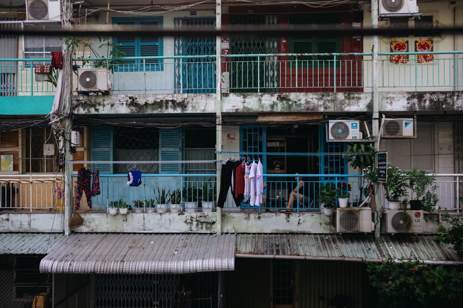 Balcón de apartamento desgastado con secadora de ropa, plantas y aires acondicionados en un entorno urbano.