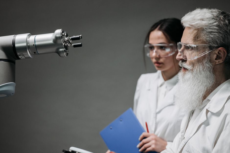 Dos científicos con batas de laboratorio y gafas analizando un brazo robótico en un entorno de laboratorio.