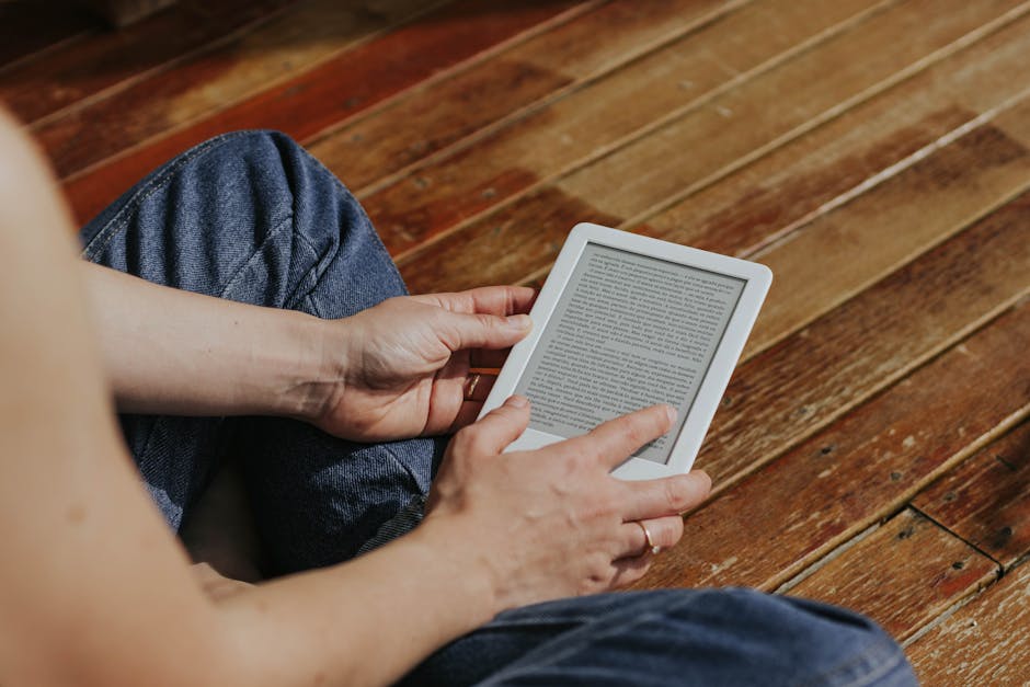 Primer plano de manos que sostienen un lector electrónico en el interior sobre un piso de madera, enfatizando la lectura digital.