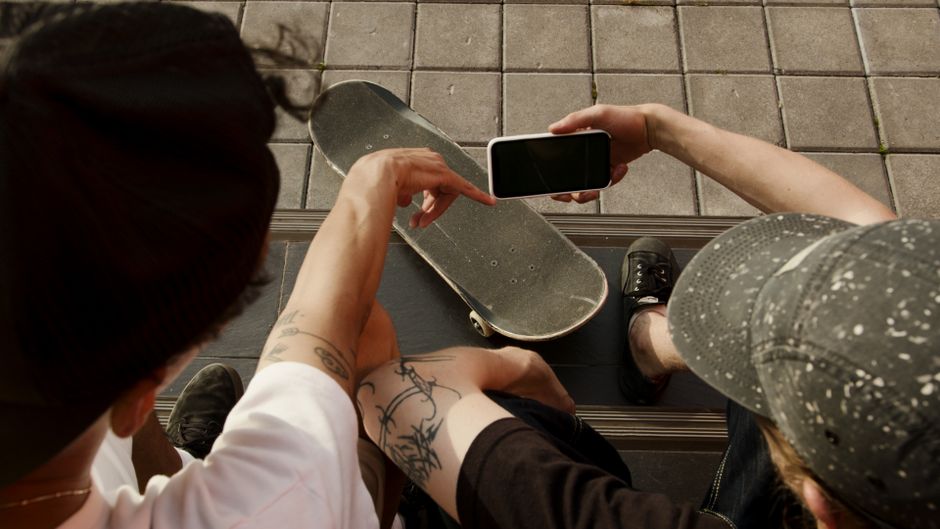 Dos adolescentes sentados al aire libre con una patineta, mirando un smartphone, disfrutando de su tiempo libre juntos.