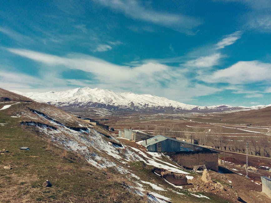 Paisaje rural pintoresco con montañas nevadas y casas dispersas bajo un cielo azul.