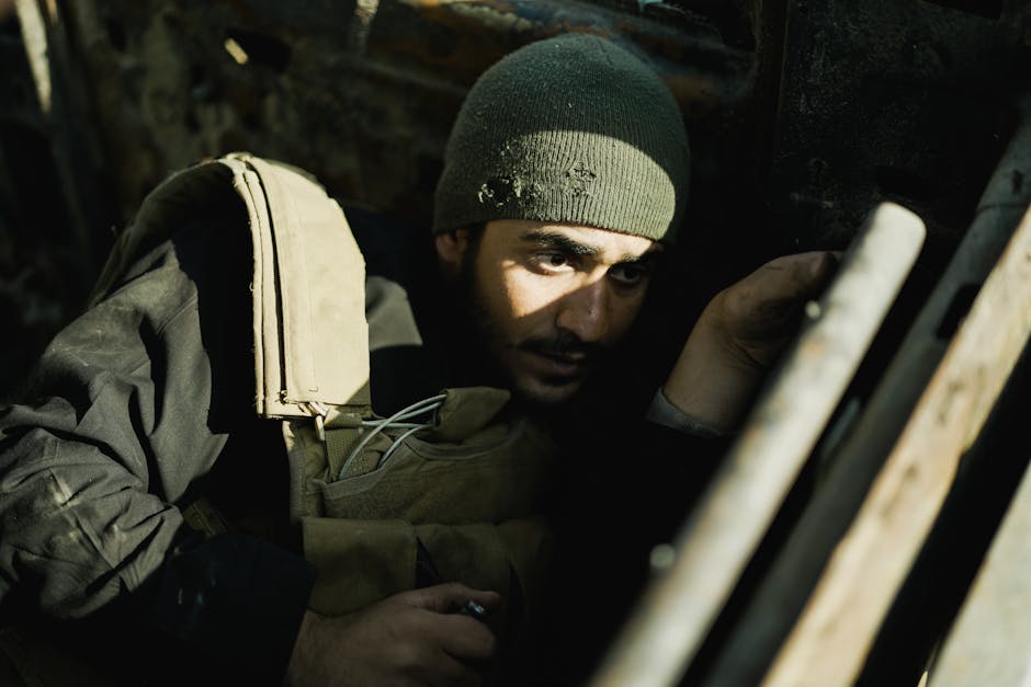 Un hombre con un gorro y un chaleco militar está sentado en un entorno con poca luz y lleno de sombras.