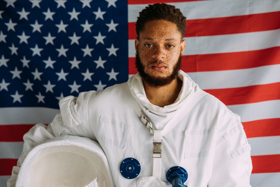 Hombre en un traje de astronauta de pie frente a un fondo de la bandera americana, mostrando un tema inspirado en el espacio.