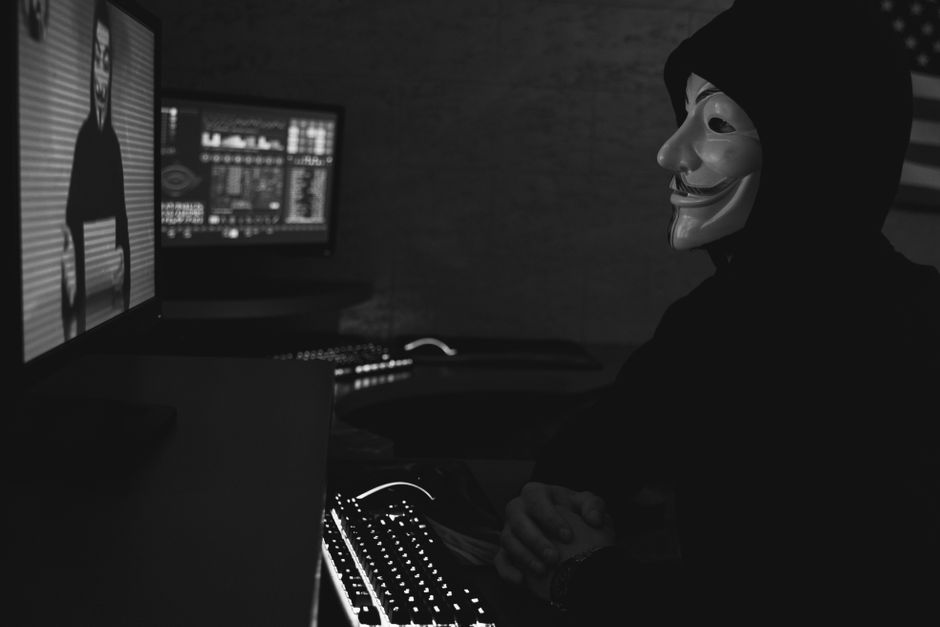 Imagen en monocromático de un hacker con una máscara de Guy Fawkes trabajando en una computadora en una habitación con poca luz.