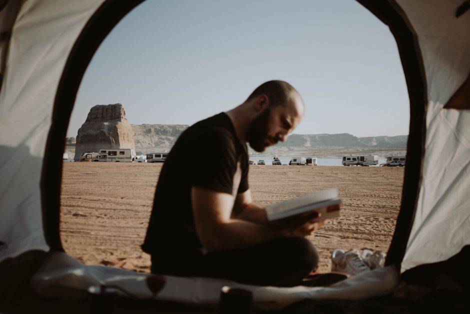 Un hombre sentado en una tienda leyendo un libro, rodeado de un impresionante paisaje desértico y autocaravanas.