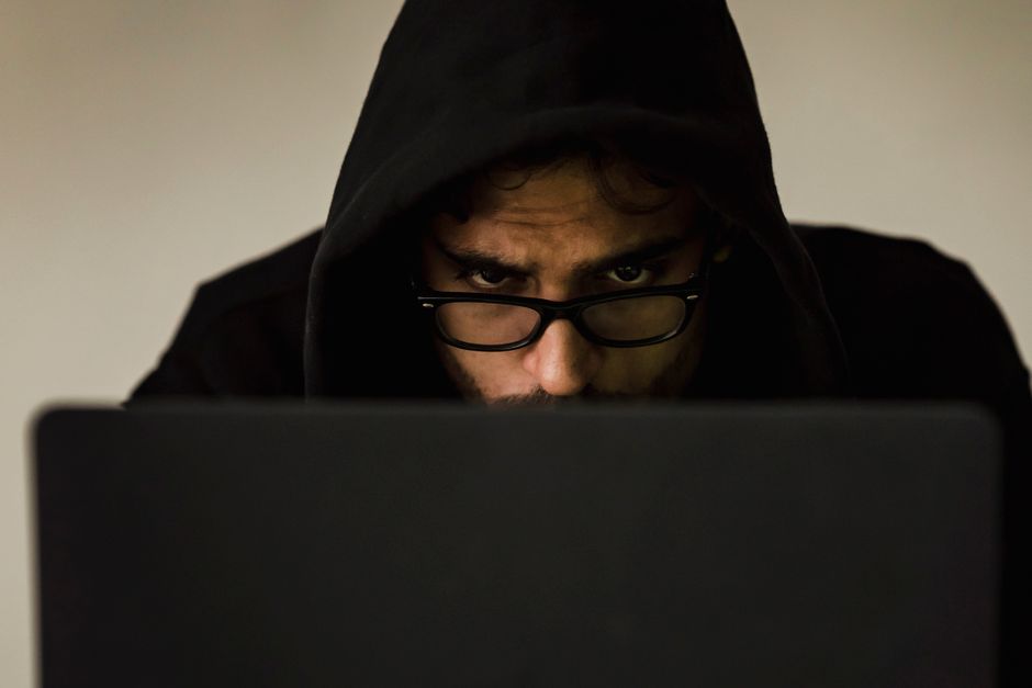 Un hacker serio con capucha centrado en la pantalla del laptop en un ambiente interno poco iluminado.