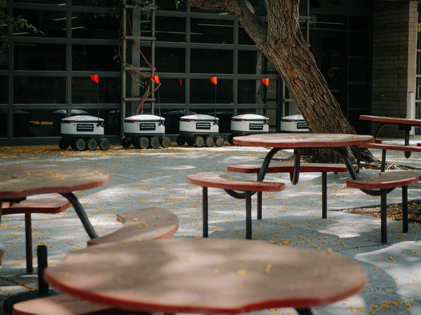 Robots de entrega futuristas alineados cerca de mesas de cafetería al aire libre, mostrando tecnología moderna.