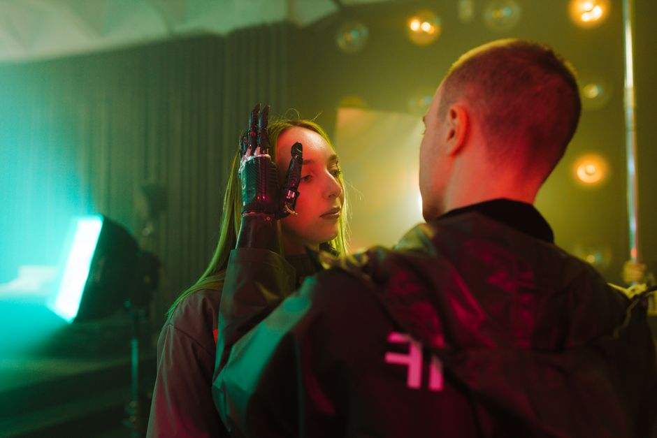 Un hombre con una mano prostética toca suavemente la cara de una mujer en un entorno cyberpunk.