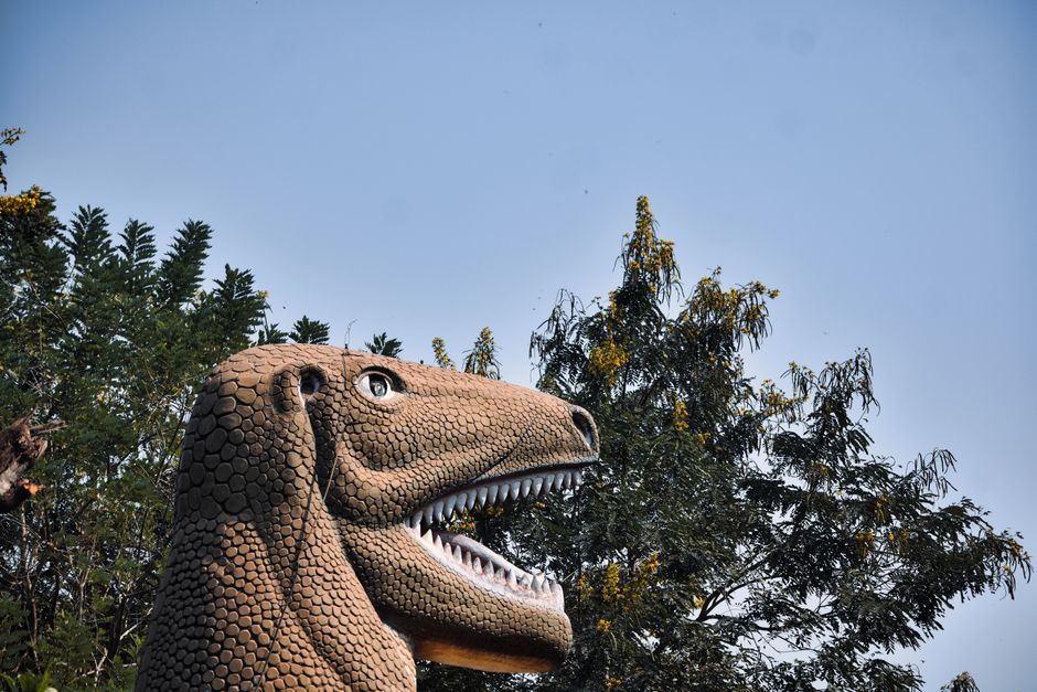Una escultura de dinosaurio realista se encuentra contra un fondo de cielo claro y árboles, creando una escena al aire libre surrealista.