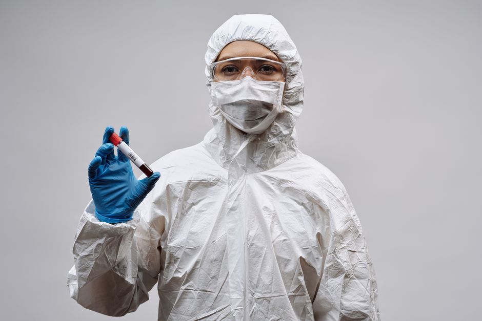Científico con equipo de protección sosteniendo una muestra de sangre en un tubo de ensayo contra un fondo gris.