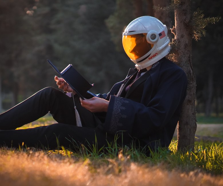Una persona que lleva un casco de astronauta lee un libro en un entorno tranquilo al aire libre.