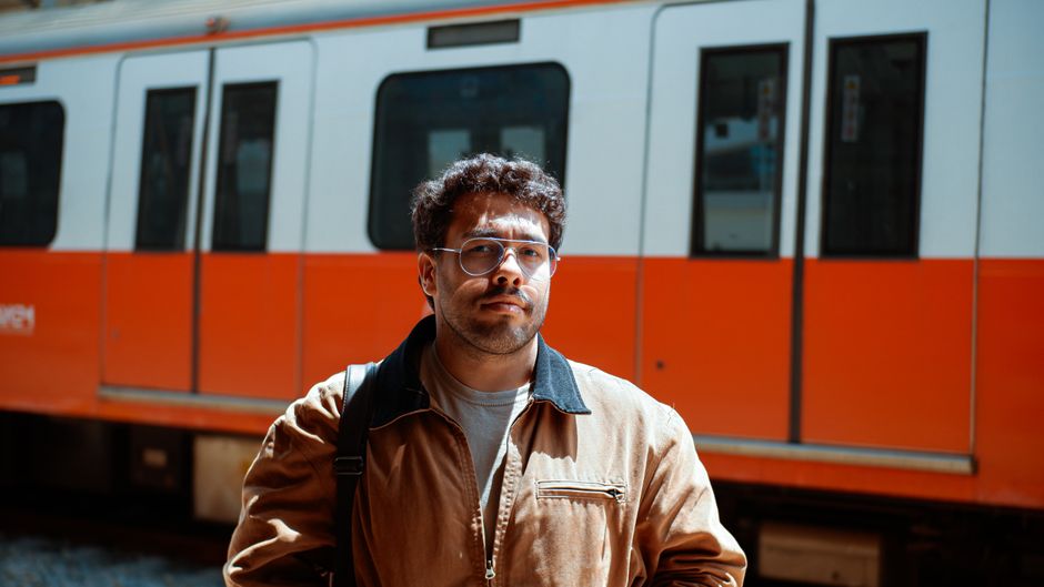 Hombre de pie en una estación de tren con un tren naranja de fondo.