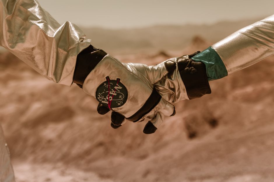 Dos astronautas en trajes brillantes tomados de la mano frente a un paisaje rocoso similar a Marte.