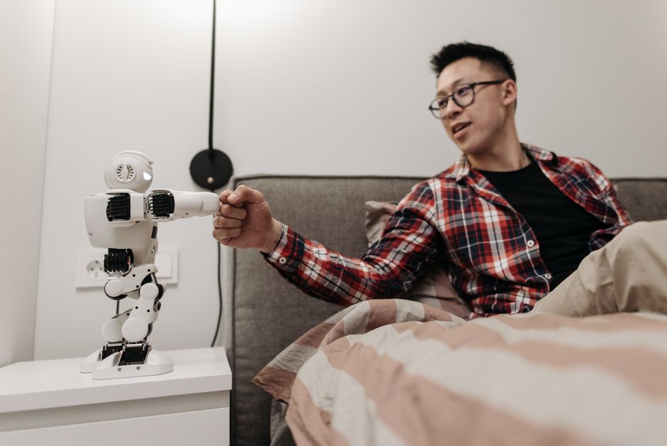 Hombre asiático sentado en la cama estrechando la mano de un robot en un dormitorio moderno.