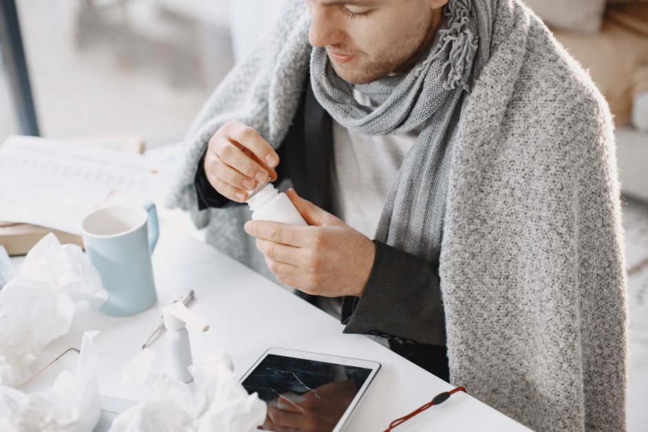 Hombre adulto con bufanda tomando medicamento en un escritorio con pañuelos y tableta, sintiéndose mal.