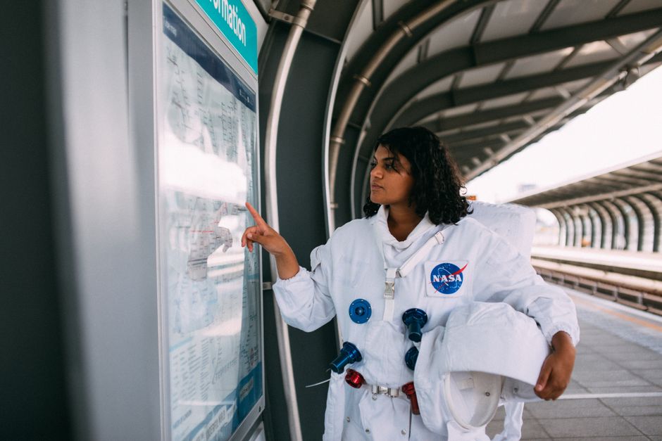 Mujer vestida con un traje de la NASA interactuando con el panel de información del metro en una estación al aire libre.