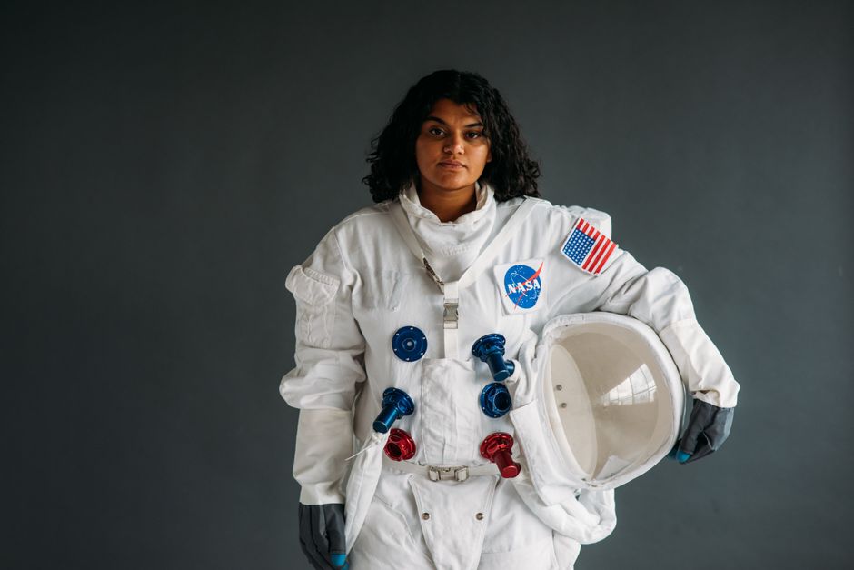 Mujer vestida de astronauta sosteniendo un casco en traje de la NASA contra un fondo gris.