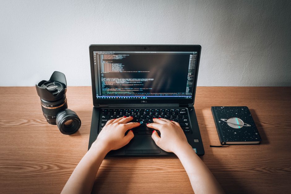 Una mujer programando en un portátil con un objetivo de cámara y un cuaderno en un escritorio de madera en una oficina en casa.