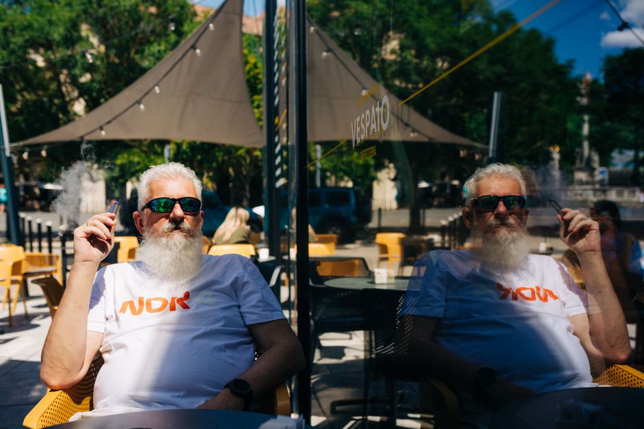 Un hombre mayor con gafas de sol disfruta de un cigarrillo en la terraza de un café, reflejado en el cristal, en un entorno urbano relajado.