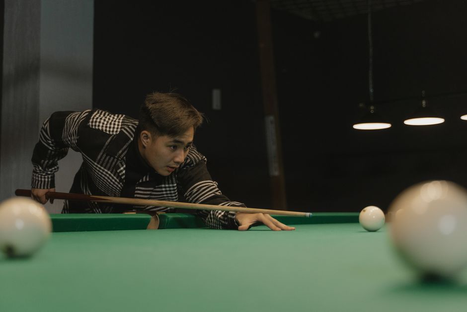 Joven apuntando con un taco en un juego de billar bajo una iluminación tenue.