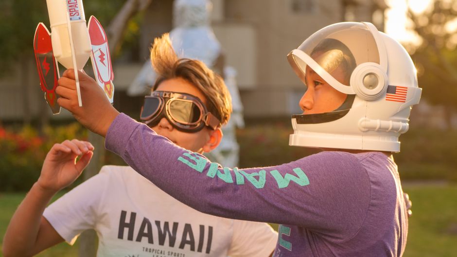 Dos niños participan en un juego imaginativo con trajes de astronauta y un juguete de cohete al aire libre.