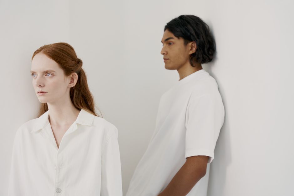 Un retrato minimalista sereno de una pareja posando contra una pared blanca, ambos con trajes blancos.